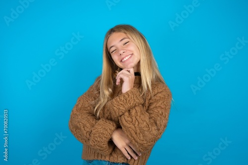 Teen caucasian girl wearing brown knitted sweater laughs happily keeps hand on chin expresses positive emotions smiles broadly has carefree expression