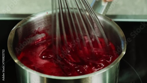 Kitchen employee stirs combination of beetroot juice in saucepan using kitchen implements. Mixture gradually begins to boil photo