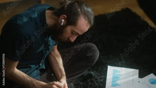 High agle shot of young adult student with long hair, wearing wireless headphones, sitting on the carpet on the floor using tablet computer writing notes while listening to music  photo