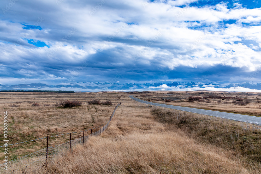 Photograph of a road heading towards a mountain range through a rural area with low level grey clouds on the South Island of New Zealand