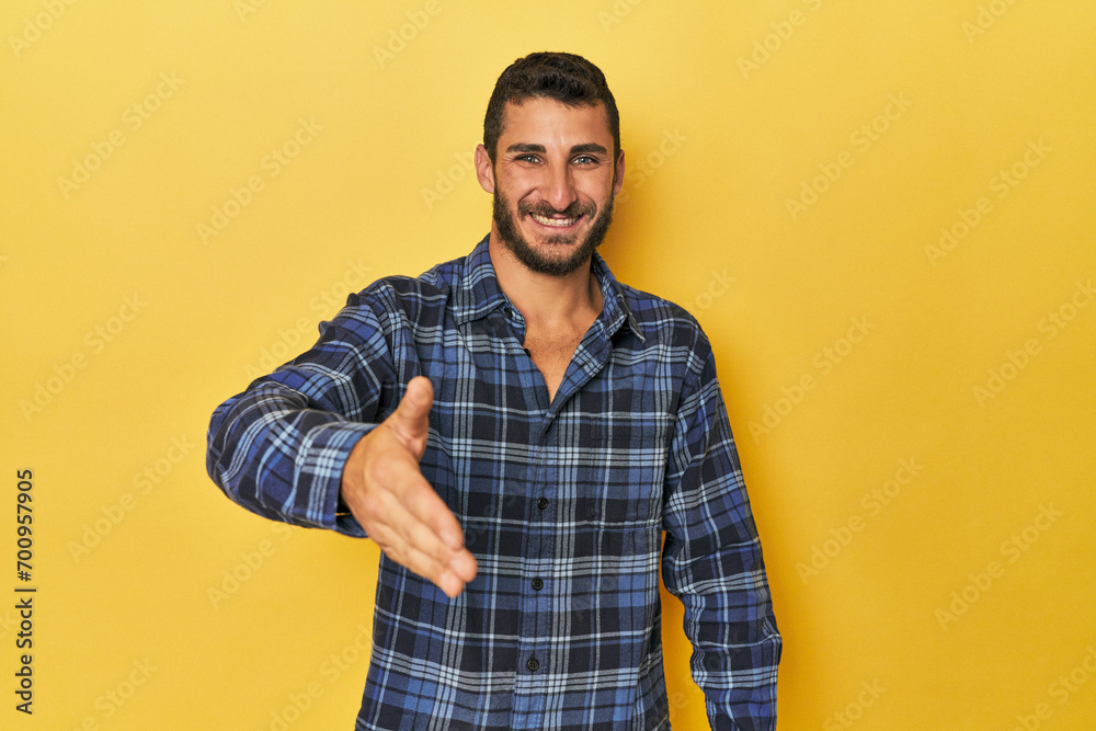 Young Hispanic man on yellow background stretching hand at camera in greeting gesture. Young Hispanic man on yellow background stretching hand at camera in greeting gesture.
