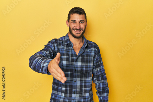 Young Hispanic man on yellow background stretching hand at camera in greeting gesture.