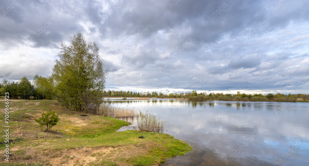 Dramatic spring landscape with clouds reflected in a pond. bank with young trees, bushes and green grass in the foreground