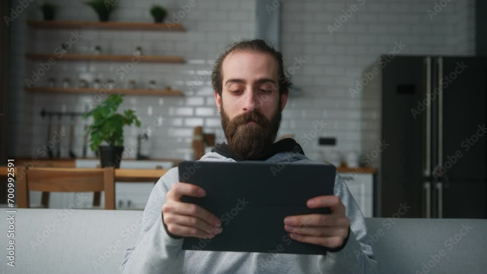 Young bearded hair bun man sitting on sofa at home with kitchen background, playing excited concentrated engrossed in interesting mobil game with tablet computer