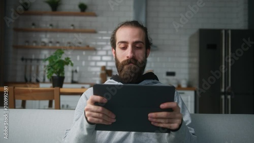 Young bearded hair bun man sitting on sofa at home with kitchen background, playing excited concentrated engrossed in interesting mobil game with tablet computer photo