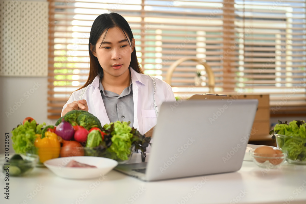 Female nutritionist in white coat calling patient and giving online consultation on laptop computer