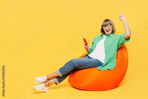 Full body elderly woman 50s year old in green shirt glasses casual clothes sit in bag chair hold in hand use mobile cell phone do winner gesture isolated on plain yellow background Lifestyle concept