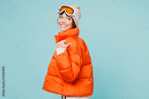 Skier young woman wear warm padded windbreaker jacket hat ski goggles mask point thumb finger back on area travel rest going spend weekend winter season in mountains isolated on plain blue background.