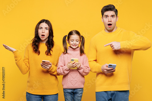 Young sad confused parents mom dad with child kid girl 7-8 years old wearing pink sweater casual clothes hold use looking at mobile cell phone isolated on plain yellow background. Family day concept.