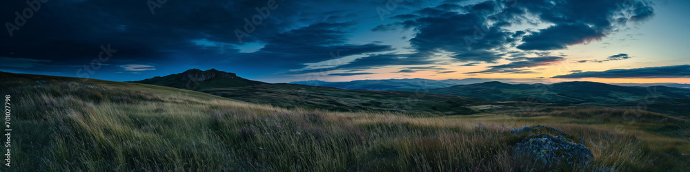 A panoramic long exposure, a grassland and meadow at evening, orange sunrise sky at far horizon, rocks on grass, dramatic blue cloudy sky, mountains and hill in background, AI generated