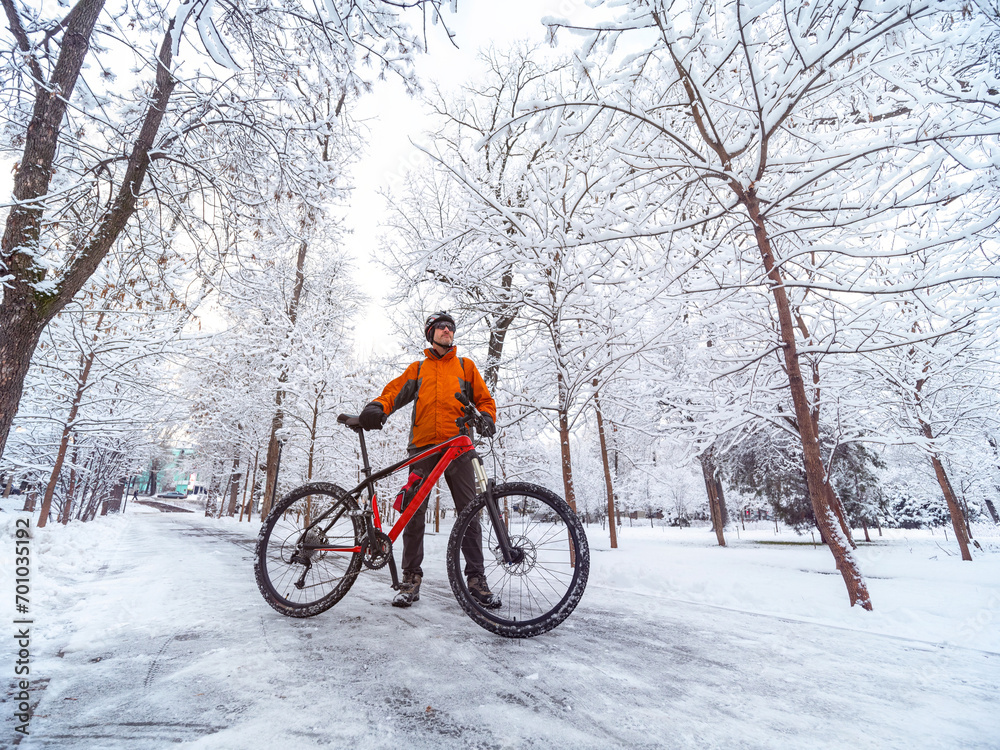 A man with a bicycle in an orange jacket and a bicycle helmet is in a winter city among snow covered trees. Active lifestyle in winter A man with a bicycle in an orange jacket and a bicycle helmet is in a winter city among snow covered trees. Active lifestyle in winter