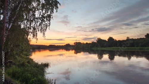On a summer evening the sun among the trees has dipped below the horizon. The colorful cloudy sky is reflected in the calm river water. Trees stand on the grassy banks, and calamus grows in the water © Balser