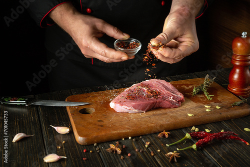 Chef adding dry spices to raw beef steak for BBQ dinner. Work environment on the kitchen table with fragrant rosemary and pepper