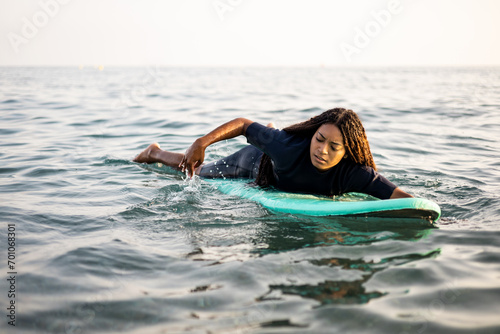 A young dark-skinned woman swimming on a surfboard in the sea dressed in a wetsuit. African women with long hair surfing. Watersports. Breaststroke swimming on a surfboard. Calm sea to practice.