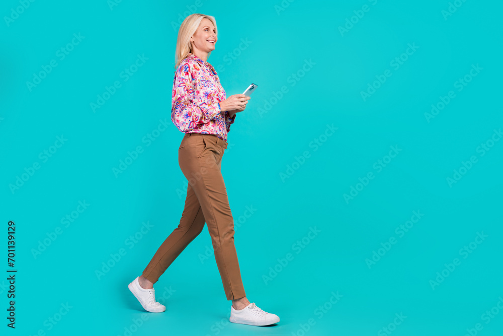 Full length photo of shiny sweet woman dressed print shirt walking typing modern device empty space isolated turquoise color background