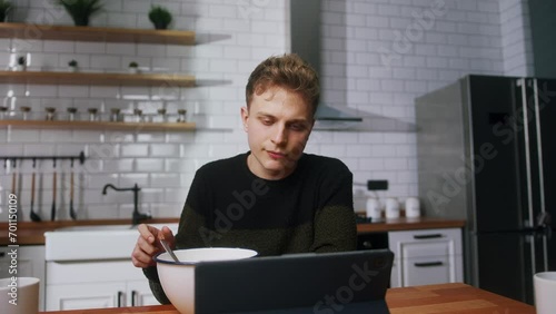 Young bored man reluctant to eat pushing a bowl full of cereal while sitting in the kitchen watching a video on a tablet computer photo