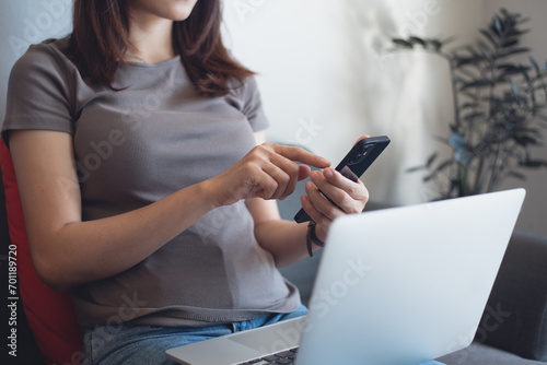 Young asian woman sitting at sofa at home, using mobile phone and working on laptop computer. Female freelancer using smartphone, surfing the internet, remote work, online job, freelance lifestye photo