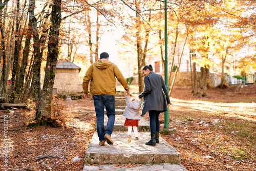 Mom and dad with a little girl walk hand in hand along a paved path in the park, lifting her above the steps