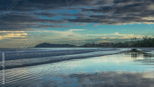 Early morning on a tropical island. The clouds in the azure sky are highlighted in golden. The ocean waves roll onto the beach. Reflection on wet shiny sand. A hill in the distance. Malaysia. Borneo.