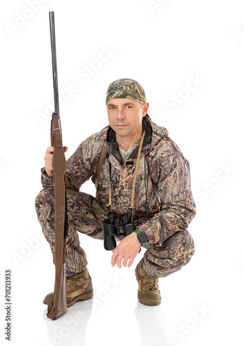 Full lengt portrait of serious duck hunter with a rifle and binoculars dropped to a crouch, isolated on white background. Fifty-year-old man in hunting uniform posind in studio.