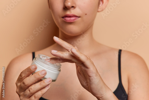 Unknown woman holds jar of nourishing cream going to apply on skin applies makeup products undergoes beauty treatments stands bare shouldered isolated over brown background. Facia care concept