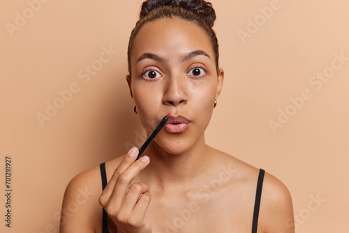 Beauty and cosmetology. Indoor photo of young pretty surprised European model standing in centre isolated on beige background looking straight holding red lip pencil near lips wearing undershirt