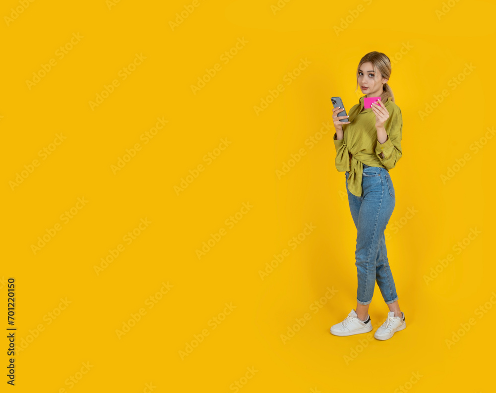 Holding mobile phone and credit card. Full body view young caucasian blonde woman standing over isolated yellow background. Online shopping, payment concept idea. Copy space. Holding mobile phone and credit card. Full body view young caucasian blonde woman standing over isolated yellow background. Online shopping, payment concept idea. Copy space.