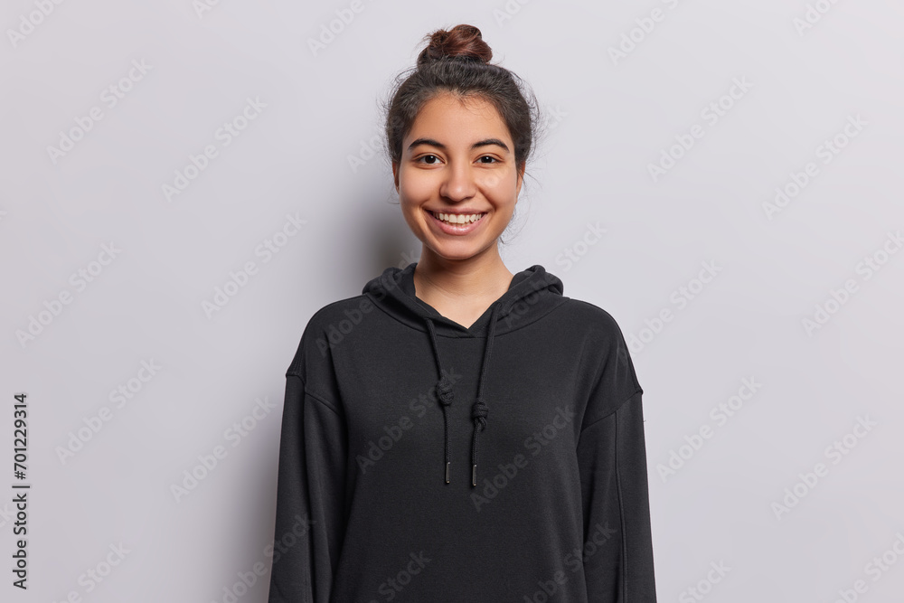 People positive emotions concept. Indoor waist up of young happy smiling Iranian girl looking straight at camera keeping hands down standing in centre isolated on white background in black hoodie People positive emotions concept. Indoor waist up of young happy smiling Iranian girl looking straight at camera keeping hands down standing in centre isolated on white background in black hoodie