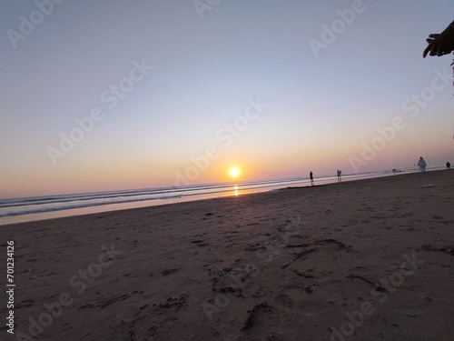 Sunset over the ocean with a sandy beach in the foreground and silhouettes of people in the distance.