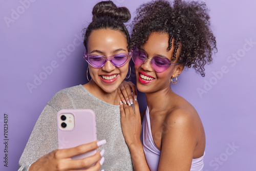 Cheerful brunette women take selfie via smartphone click photos on front camera wear sunglasses and stylish clothing isolated over purple wall spending time together filming content for her video blog