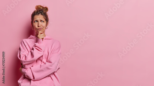 Horizontal shot of contemplative thoughtful European woman keeps hand on chin has expression of intrigue and uncertainty purses lips dressed in casual hoodie isolated over pink background copy space photo