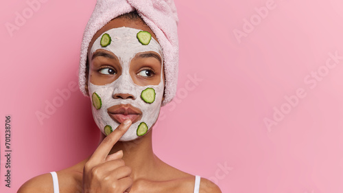 Beauty and cosmetology. Studio shot of young dreamy relaxed European girl applying clay face mask and slices of cucumber standing isolated on left on pink background looking at space for promotion