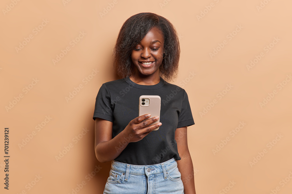 Smiling dark skinned woman using smartphone app chatting or ordering holding mobile phone reads pleasant message has happy smile on face dressed in black t shirt and jeans isolated over brown wall Smiling dark skinned woman using smartphone app chatting or ordering holding mobile phone reads pleasant message has happy smile on face dressed in black t shirt and jeans isolated over brown wall