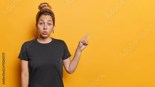 Special advert offer. Stunned young European woman with hair gathered in bun points index finger on copy space feels impressed wears black t shirt isolated over yellow background. Shocking offer