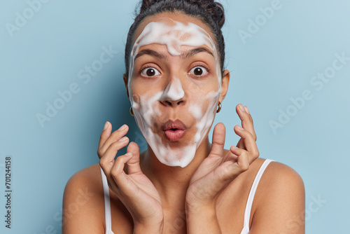 Horizontal shot of impressed Latin woman washes face with white soap cleans pores keeps lips folded feels stunned isolated over blue background. Skin care pampering and daily hygiene concept photo