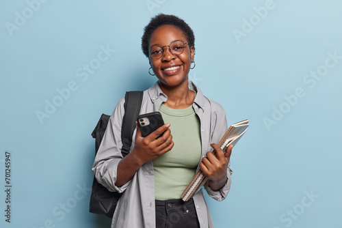 Foto Horizontal shot of pretty dark skinned female teenager holds mobile phone and notebooks being in good mood studies at college or university carries backpack isolated over blue studio background
