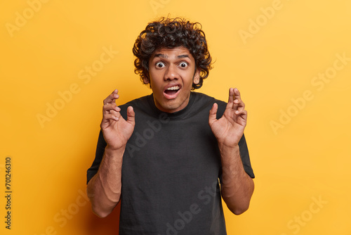 Studio shot of young scared Hindu male student wearing casual black tshirt standing isolated in centre on yellow background seeing something unexpected or unpleasant expressing negative emotions