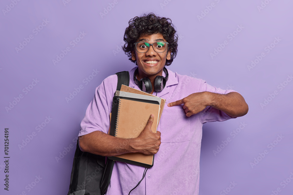 People positive emotions concept. Indoor photo of young happy smiling Hindu male student standing in centre isolated on purple background looking straight at camera holding books with headphones