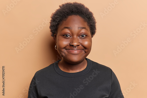Portrait of cheerful dark skinned chubby woman smiles pleasantly has piercing in nose glad to talk with friend wears casual black t shirt isolated over brown background. People and emotions concept © Wayhome Studio