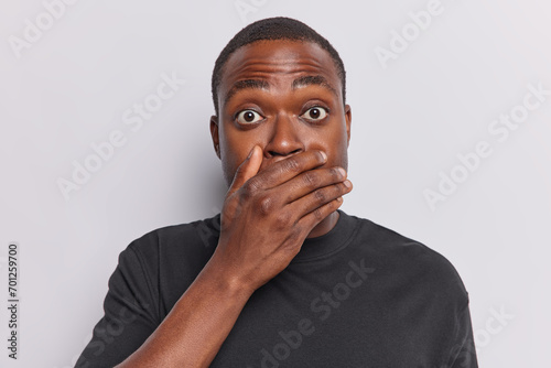 Secret concept. Dark skinned young man covers mouth with hand tries to be speechless has bugged eyes dressed in black t shirt isolated over white studio background standing intense and scared photo