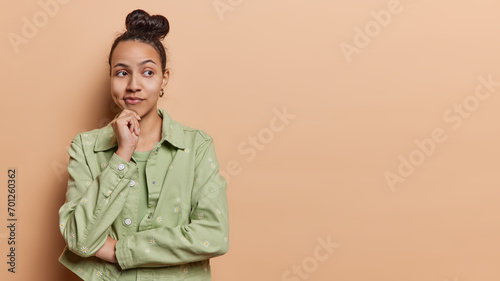 Fotografie Thoughtful young Latin woman keeps hand on chin concentrated aside has dimple on cheek wears green jacket being deep in thoughts isolated over brown background copy space for your promotion