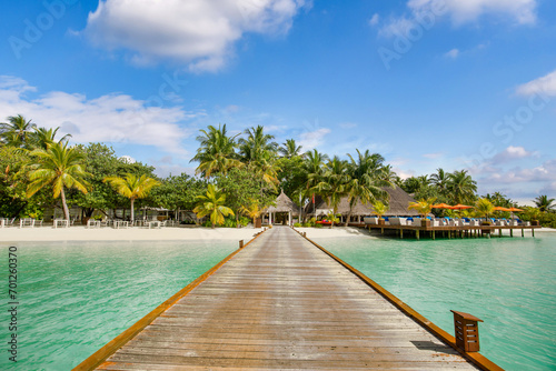 Luxury travel landscape. Water villas, wooden pier bridge leads to palm trees over white sandy shore close to blue sea, seascape. Summer panoramic vacation, beach resort on tropical island paradise 