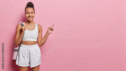 Horizontal shot of satisfied Latin woman dressed in white cropped top and shorts carries rolled karemat on shoulder indicates index finger on copy space for your advertisement isolated over pink wall