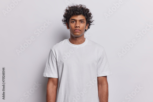 Waist up shot of serious curly haired Hindu man looks directly at camera keeps arms down has confident expression dressed in casual t shirt isolated on white background. Human face expressions concept © wayhome.studio 