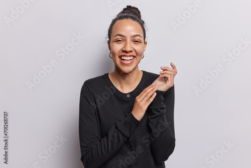 Horizontal shot of cheerful pretty Latin woman rubs palms and laughs joyfully hears funny joke from friend expresses positive sincere emotions wears black jumper isolated over white background photo