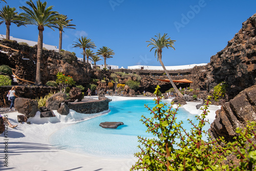 Pool of blue waters and palemeeras, outside the cave of Los Jameos del Agua. Light at the end of the cave. Sky with big white clouds. Lanzarote, Canary Islands, Spain.