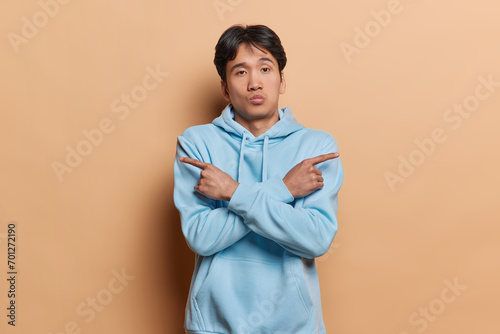 Waist up shot of hesitant Japanese man points at different sides as if choosing between two options feels doubtful poses against brown background tries to make difficult choice. This or that