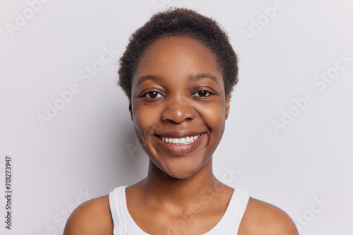 Portrait of lovely dark skinned pretty teenage girl with short curly hair smiles toothly has perfect even teeth dressed in casual t shirt isolated over white background feels confident in herself