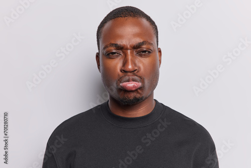 Troubled offended dark skinned man has sulking expression purses lips feels unhappy has some problems in life being hurt by bad words dressed in casual basic black t shirt isolated on white background photo