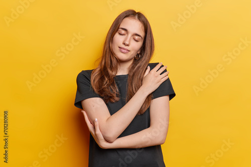 Selfish long haired woman with closed eyes embracing herself tenderly daydreams about something wears casual black t shirt isolated over yellow wall. Romantic female model recalls nice lovely memories photo